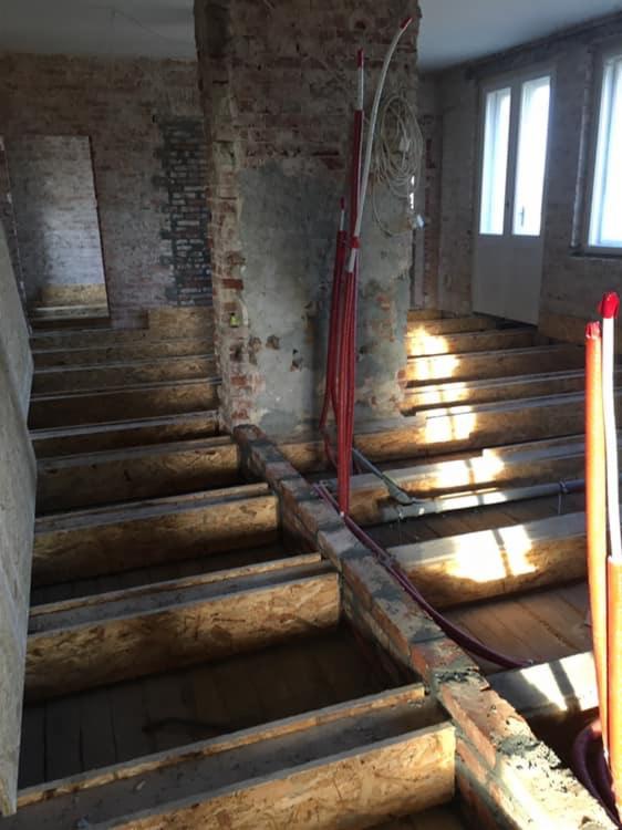 Wnętrze budynku w trakcie remontu, widoczne odsłonięte ceglane ściany, drewniane legary pod podłogę i czerwone rury instalacyjne.