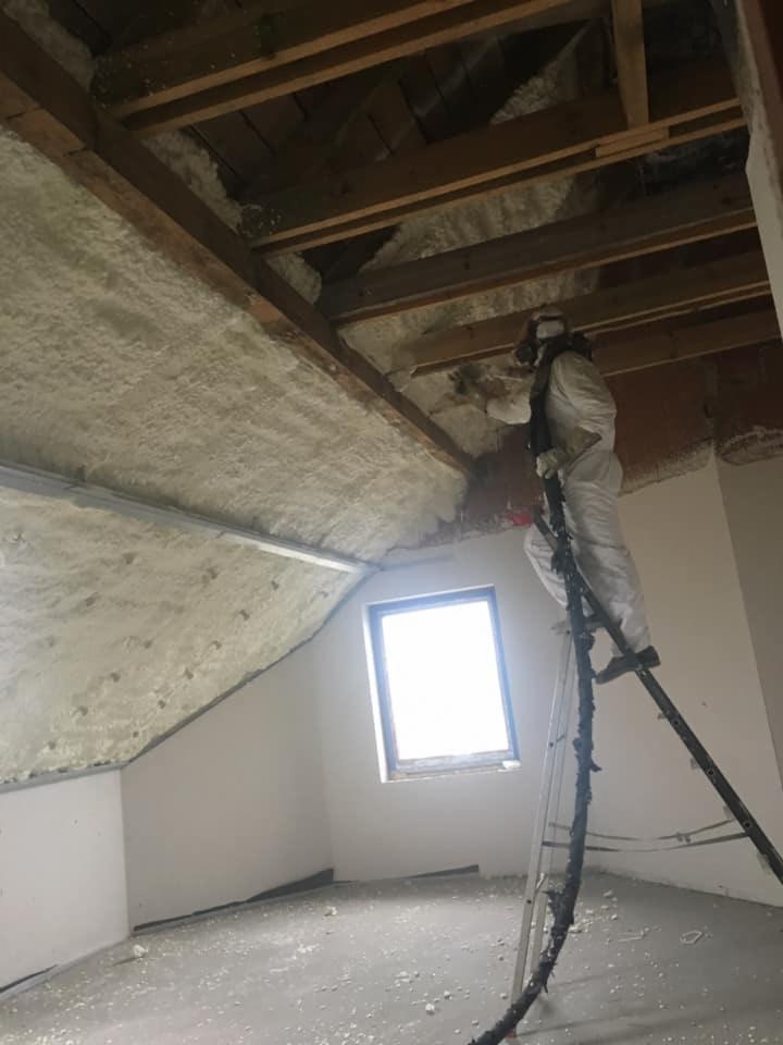 Pracownik w kombinezonie ochronnym na drabinie aplikuje piankę PUR na poddaszu z widoczną konstrukcją drewnianą i oknem.