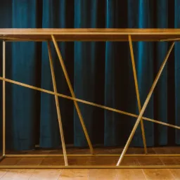 Konsola z blatem z jasnego drewna i geometryczną, złotą podstawą na tle ciemnoturkusowych zasłon.