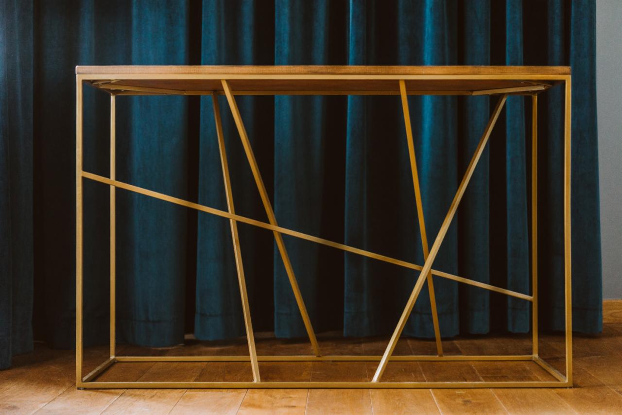 Konsola z blatem z jasnego drewna i geometryczną, złotą podstawą na tle ciemnoturkusowych zasłon.