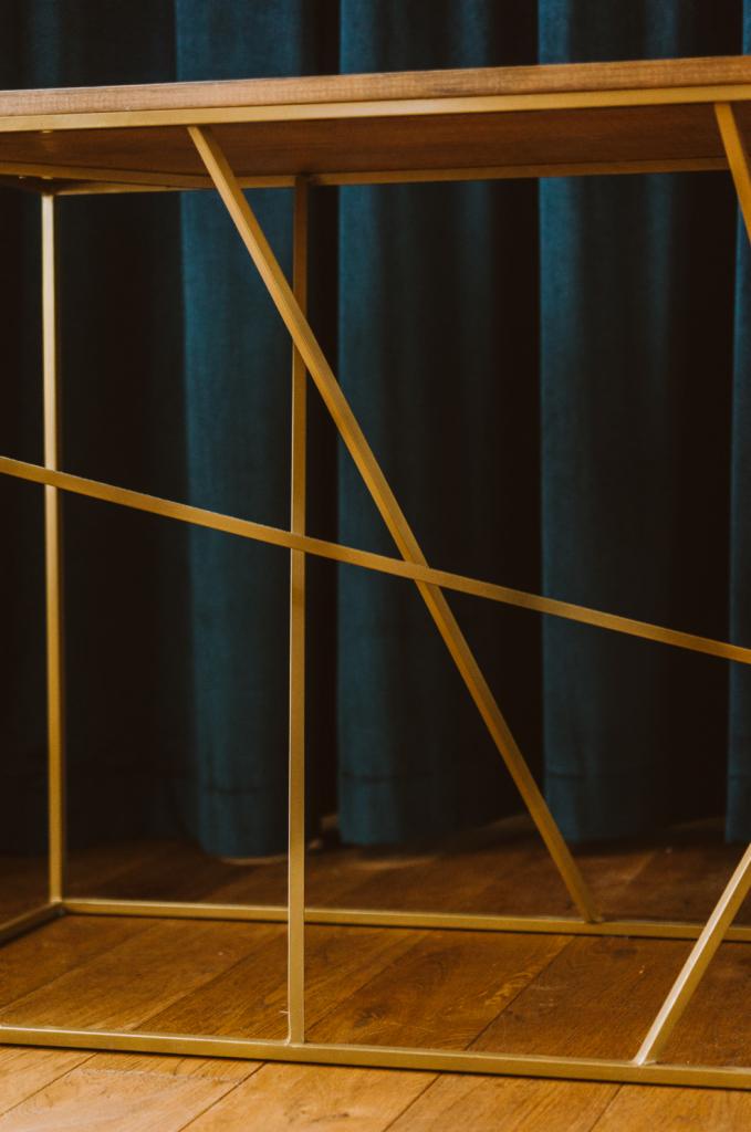 Stolik z blatem drewnianym i geometryczną, złotą podstawą na tle ciemnoturkusowych zasłon.