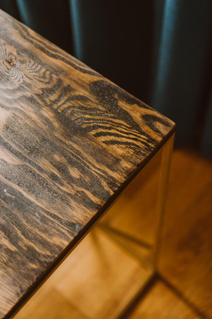 Fragment blatu drewnianego o wyraźnym usłojeniu, wykończonego ciemnym bejcowaniem na metalowej podstawie, na tle ciemnej zasłony.