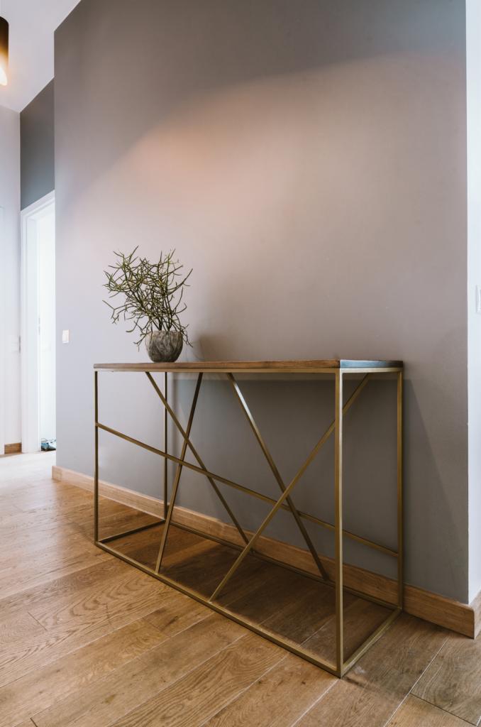 Nowoczesna konsola z drewnianym blatem i geometryczną, złotą podstawą, na tle szarej ściany i drewnianej podłogi, z dekoracyjną rośliną w ceramicznej doniczce.