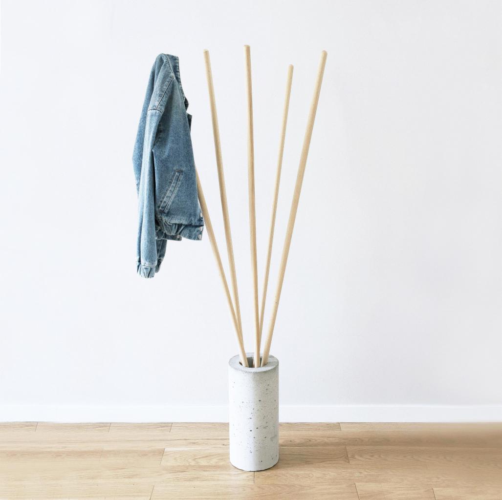 Minimalistyczny wieszak na ubrania: jasne drewniane pręty w betonowej podstawie, wisząca jeansowa kurtka na tle białej ściany i drewnianej podłogi.