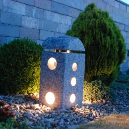 Granitowa lampa ogrodowa z okrągłymi otworami, oświetlona ciepłym światłem, otoczona zielenią i kamieniami, na tle muru z szarego kamienia.