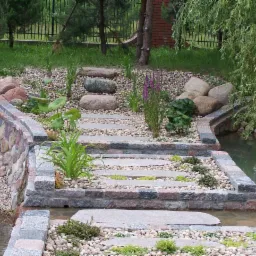 Kamienne schody prowadzące przez strumień w ogrodzie, otoczone roślinnością i ozdobnymi kamieniami.