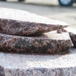 Surowe, obrobione fragmenty czerwono-szarego granitu z widoczną strukturą kamienia, ułożone warstwowo, z kołem samochodu w tle.