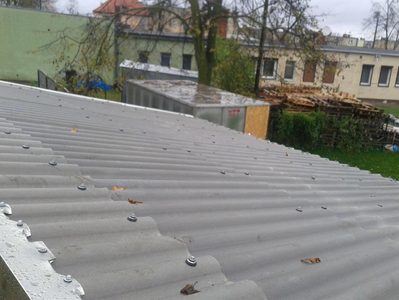 Szary dach z falistej blachy, mocowany śrubami z uszczelkami, widoczne krople deszczu i otoczenie z budynkami i paletami w tle.