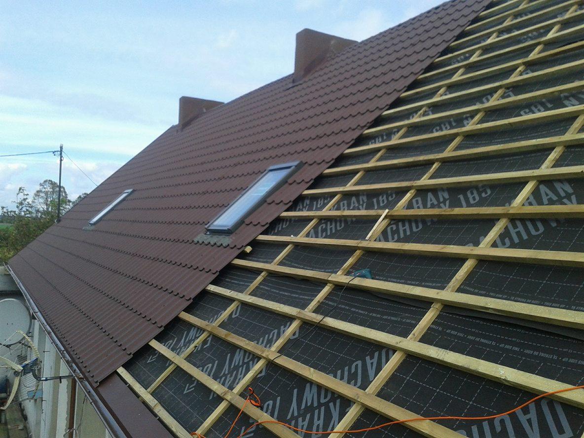 Częściowo pokryty brązową blachodachówką dach z oknami połaciowymi, widoczne drewniane łaty i membrana dachowa na niezadaszonej części.