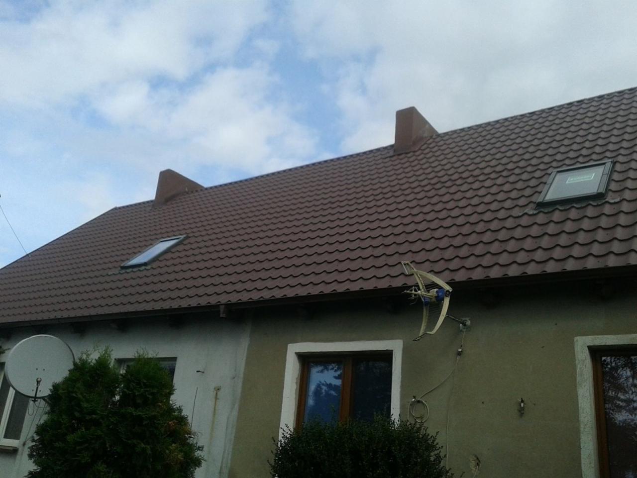 Brązowy dach z dachówki trapezowej z dwoma oknami dachowymi i kominem na budynku z anteną satelitarną na elewacji.