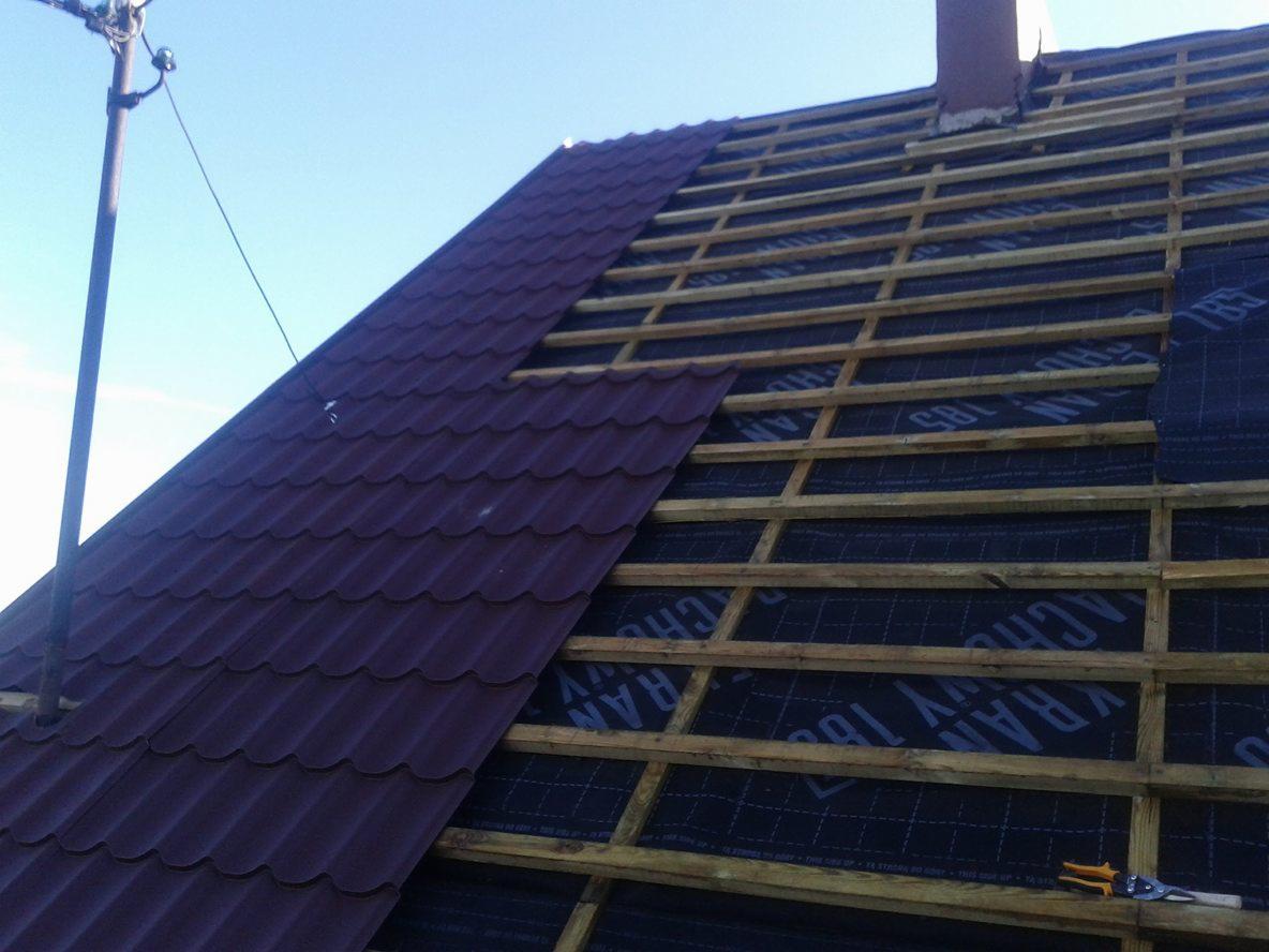 Połowa dachu pokryta bordową blachodachówką, druga połowa z widoczną konstrukcją drewnianą i membraną dachową, na dole nożyce do blachy.