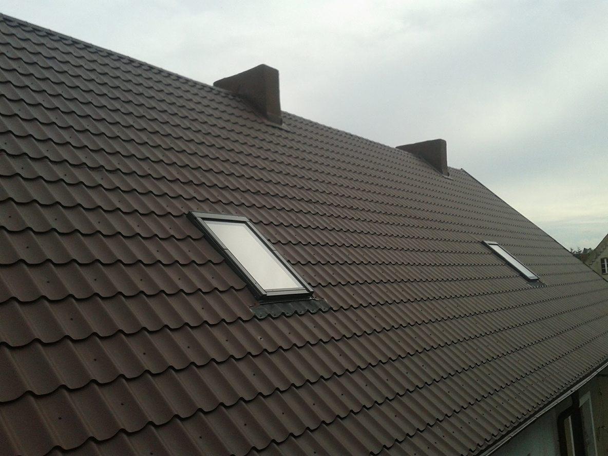 Brązowy dach z blachy trapezowej z dwoma oknami dachowymi i trzema kominami wentylacyjnymi na tle pochmurnego nieba.