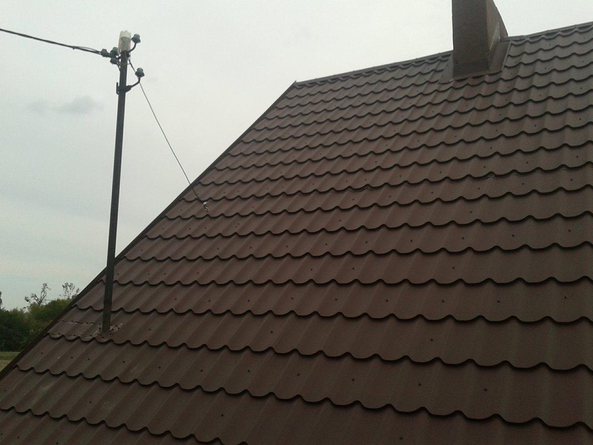 Brązowy dach z blachy trapezowej z kominem i masztem antenowym na tle pochmurnego nieba.