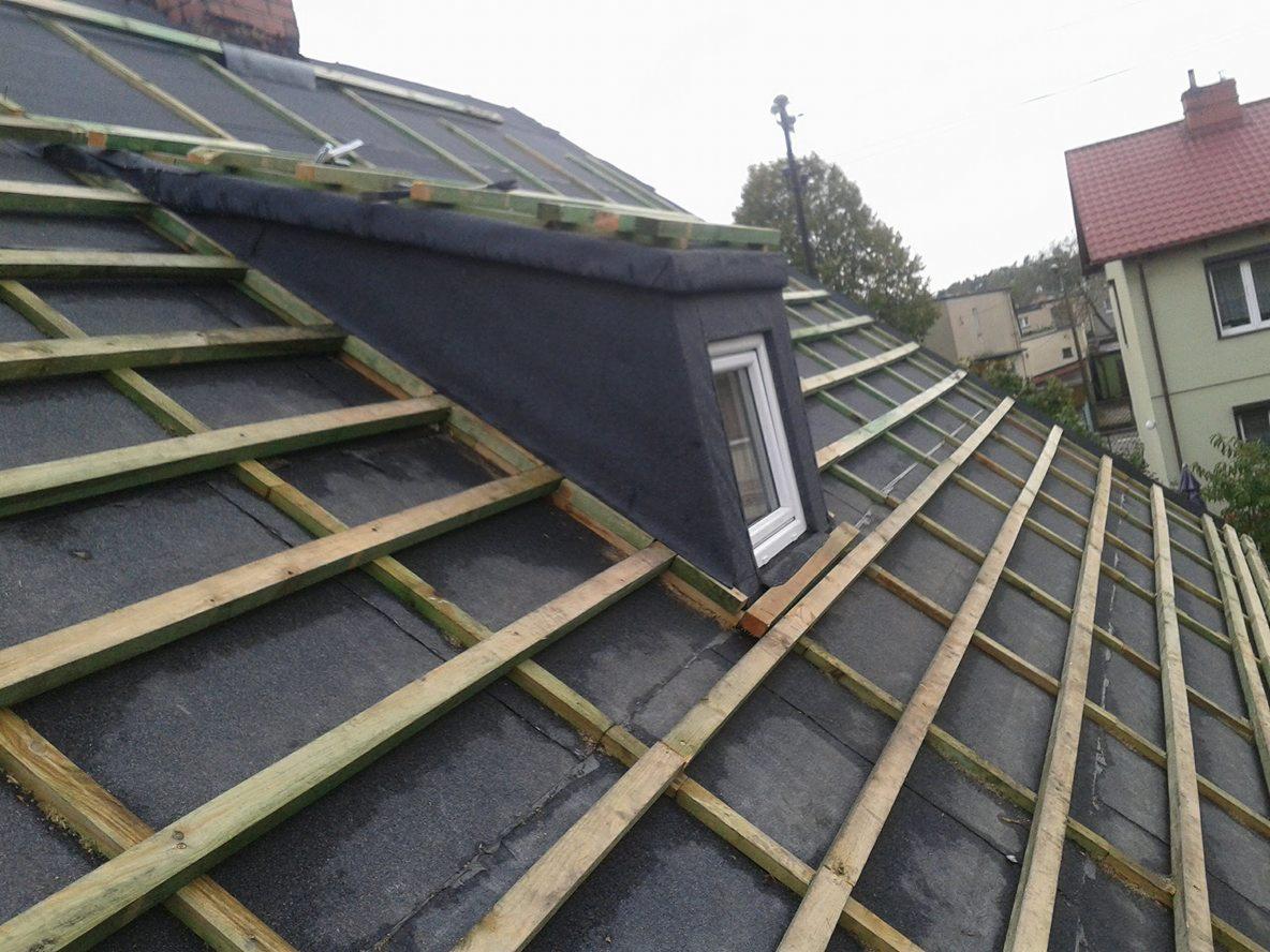 Dach w trakcie budowy z widocznym oknem dachowym i konstrukcją drewnianą, pokryty czarną membraną dachową, w tle budynki mieszkalne.