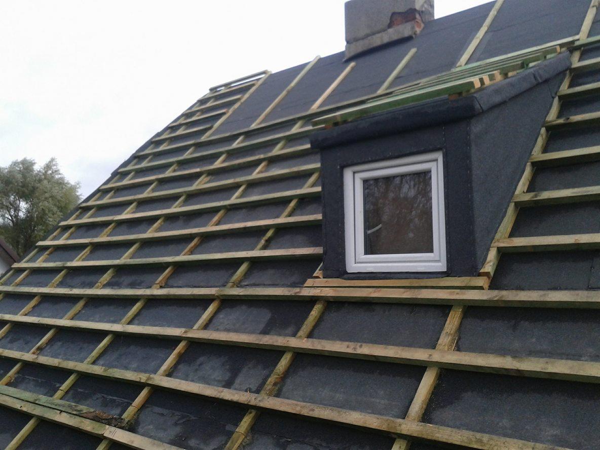 Dach w trakcie krycia z widoczną konstrukcją drewnianą, papą i oknem dachowym, z fragmentem komina w tle.