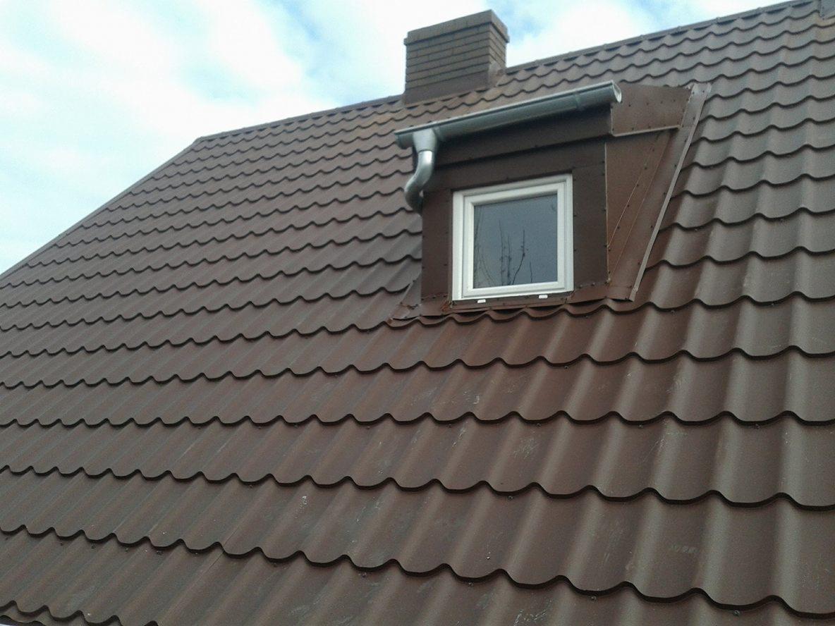 Brązowy dach z dachówki ceramicznej z oknem dachowym, orynnowaniem i kominem w tle, widziany pod kątem.
