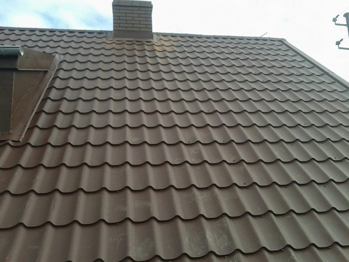 Brązowy dach z blachodachówki falistej, widoczny komin z cegły i fragment rynny, ujęcie z dołu.