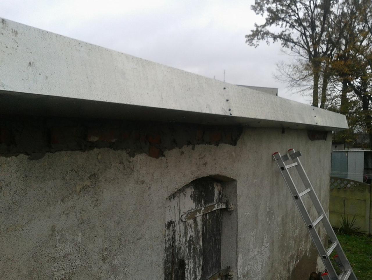Szary betonowy daszek nad starą, otynkowaną budowlą z widocznymi cegłami, drabina oparta o ścianę, pochmurne niebo w tle.