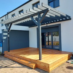 Krzysztof Mikołajczak - Nowoczesny taras drewniany z pergolą w kolorze antracytowym, przylegający do białego domu z szarymi akcentami. Widoczne donice z kwiatami i trawnik.