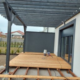 Krzysztof Mikołajczak - Drewniany taras w trakcie budowy z szarą pergolą, widoczne legary i częściowo ułożona deska tarasowa. Na tarasie narzędzia i wiaderka. Tło stanowi dom z szarymi elementami.