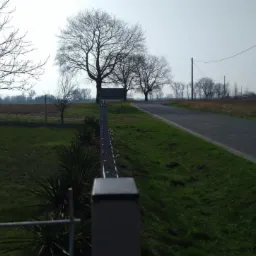 Metalowe ogrodzenie panelowe w perspektywie, oddzielające posesję od drogi w wiejskim krajobrazie, widoczne drzewa bez liści i budynek w tle.