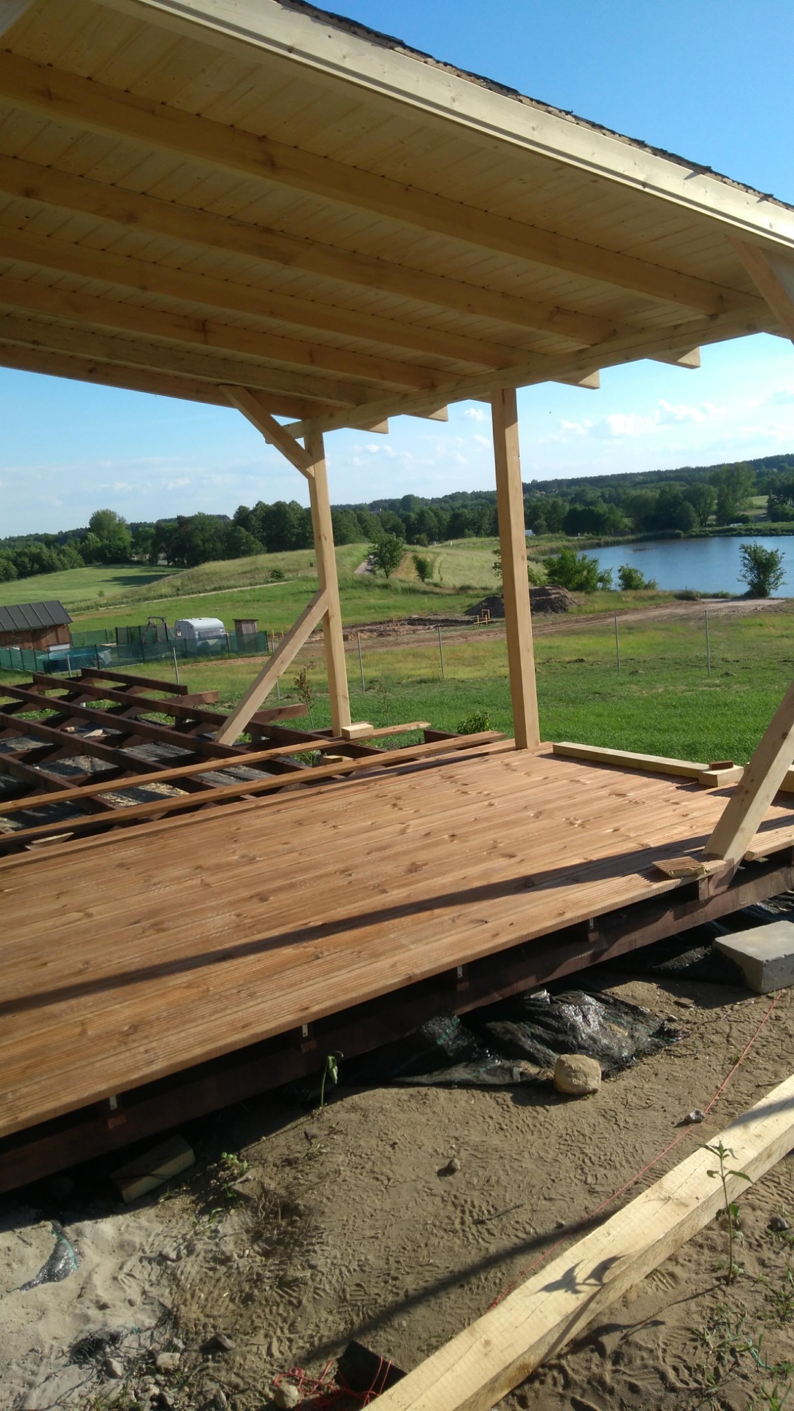 Drewniany taras w trakcie budowy z widokiem na jezioro i zielone tereny. Częściowo ułożone deski, widoczne elementy konstrukcyjne i podłoże. Jasne drewno, słoneczny dzień.
