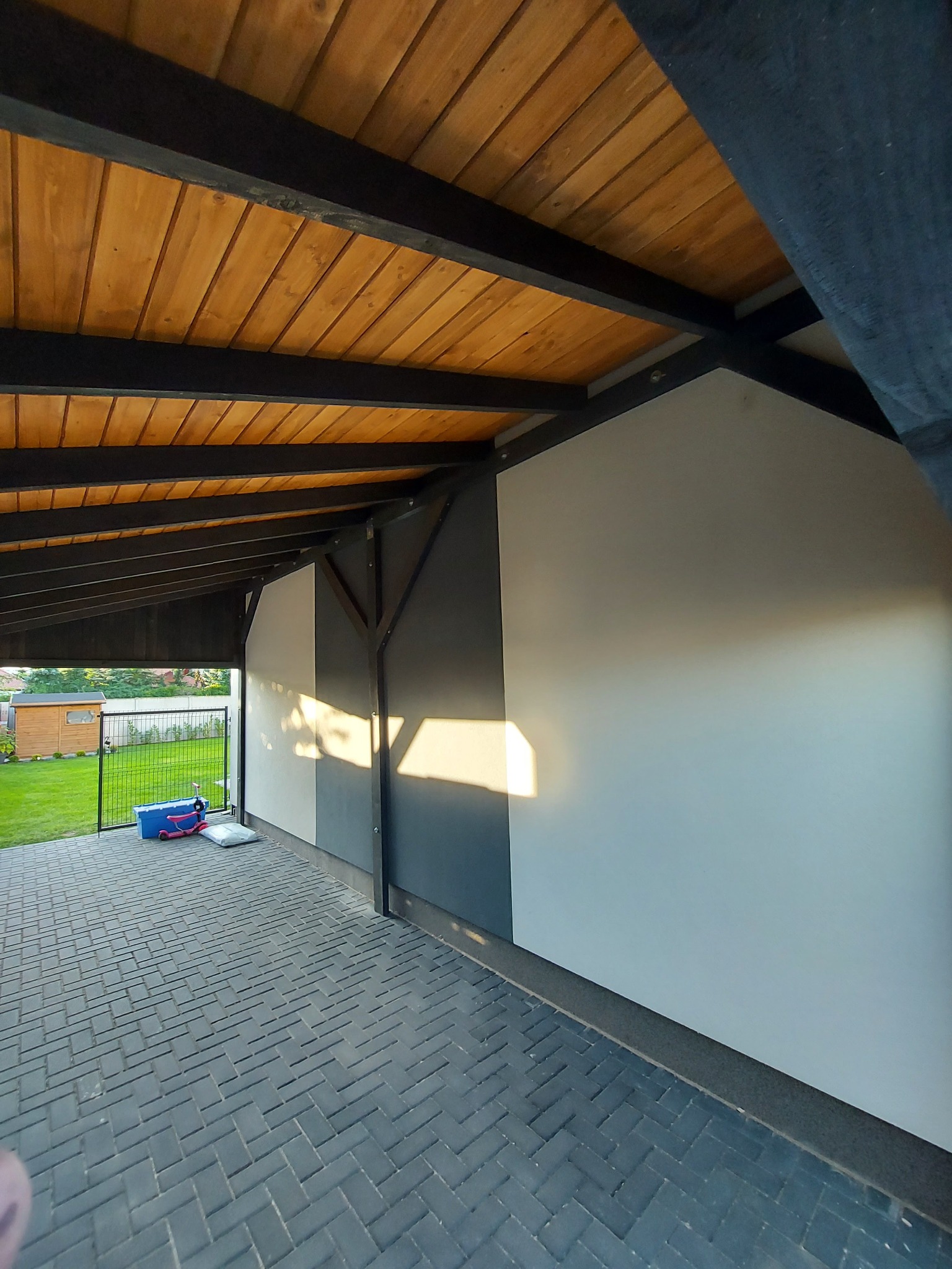 Zadaszenie tarasu z drewnianą podsufitką i szarą ścianą boczną. Na zewnątrz widoczny trawnik i mały budynek gospodarczy. Kostka brukowa na podłożu.