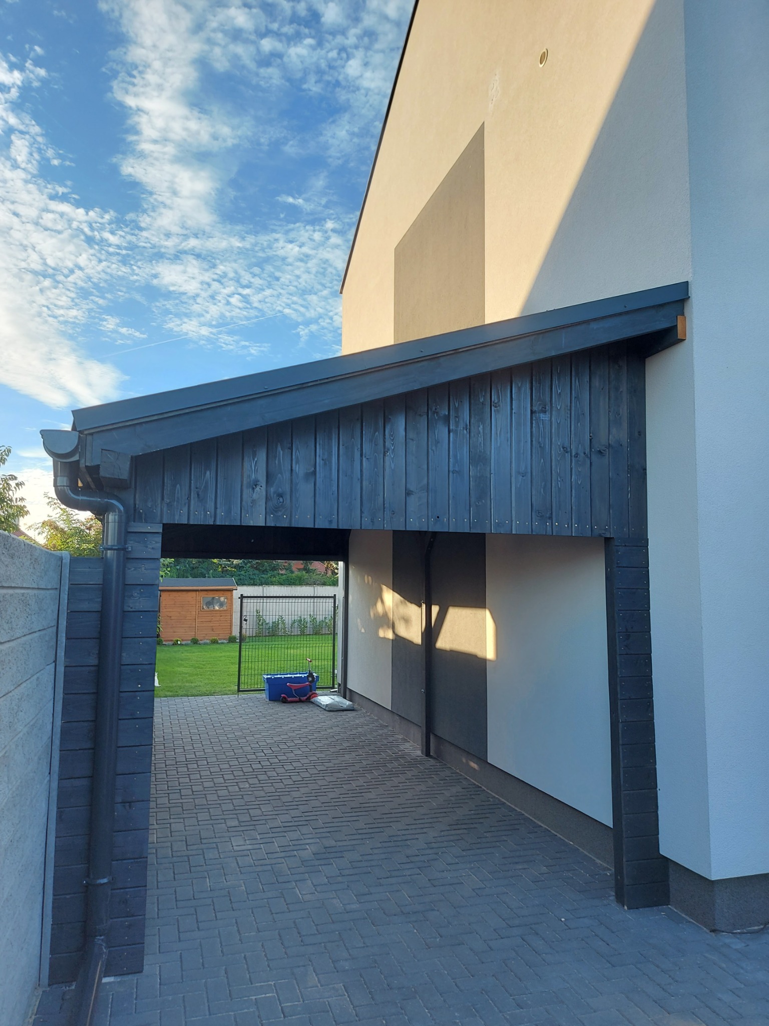 Zadaszenie nad wjazdem na posesję, wykonane z ciemnego drewna, z widoczną kostką brukową i fragmentem ogrodzenia. W tle trawnik i domek narzędziowy.