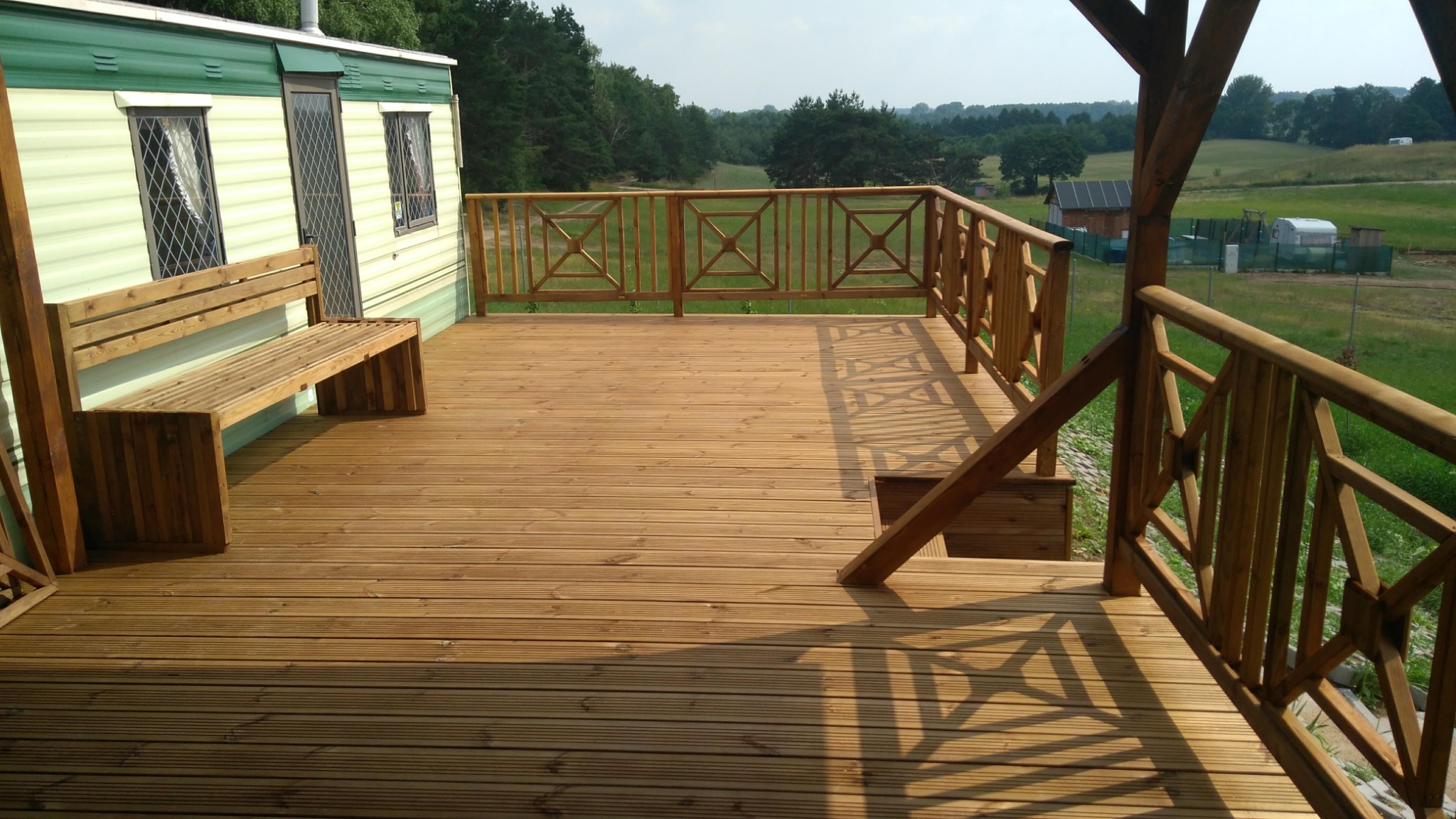 Drewniany taras z balustradą i ławką przy domku mobilnym, widok na zielone wzgórza. Geometryczny wzór balustrady rzuca cień na deski tarasu.