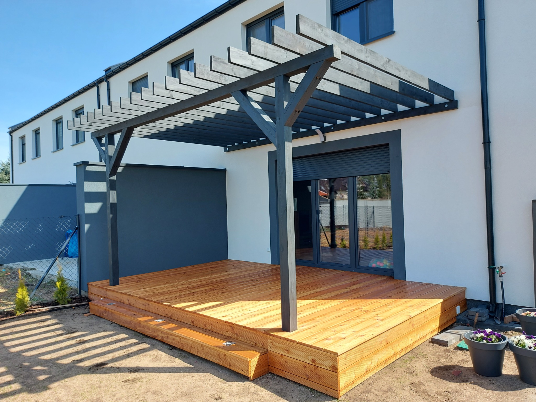 Nowoczesny taras drewniany z pergolą w kolorze antracytowym, przylegający do białego domu z szarymi akcentami. Widoczne donice z kwiatami i trawnik.