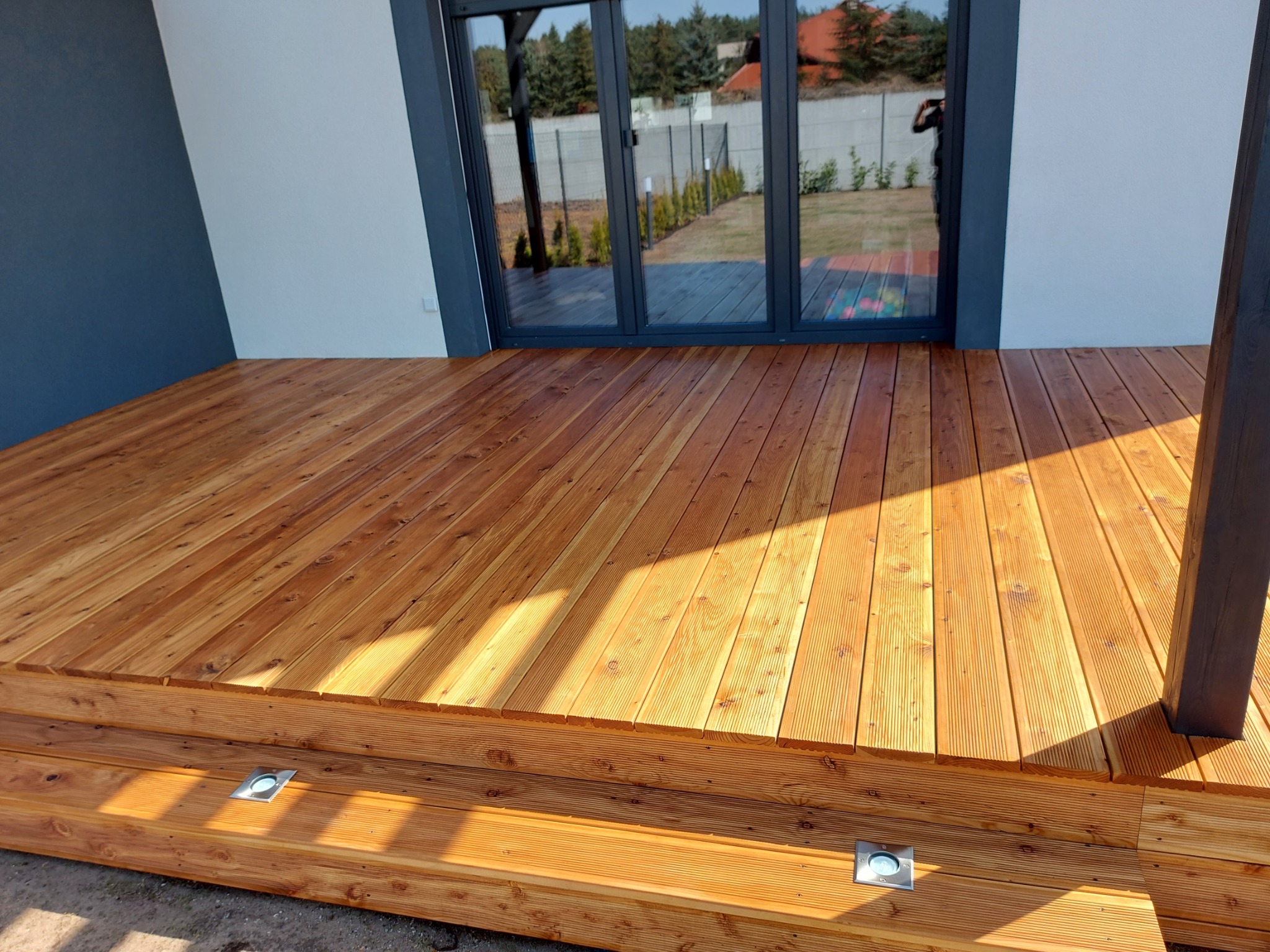 Nowy taras drewniany z oświetleniem punktowym, przylegający do domu z dużymi oknami tarasowymi. Widoczne deski tarasowe i elementy konstrukcyjne.