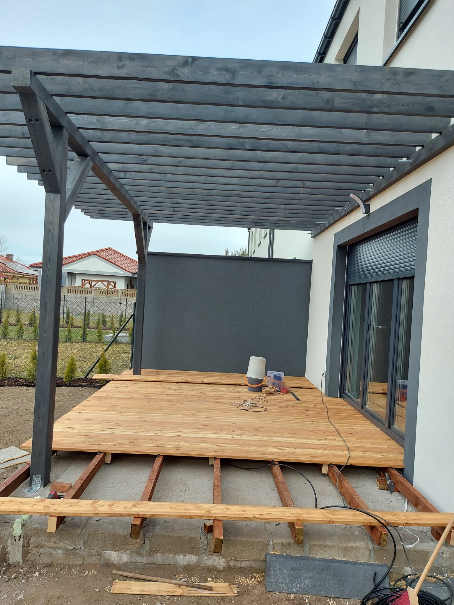Drewniany taras w trakcie budowy z szarą pergolą, widoczne legary i częściowo ułożona deska tarasowa. Na tarasie narzędzia i wiaderka. Tło stanowi dom z szarymi elementami.