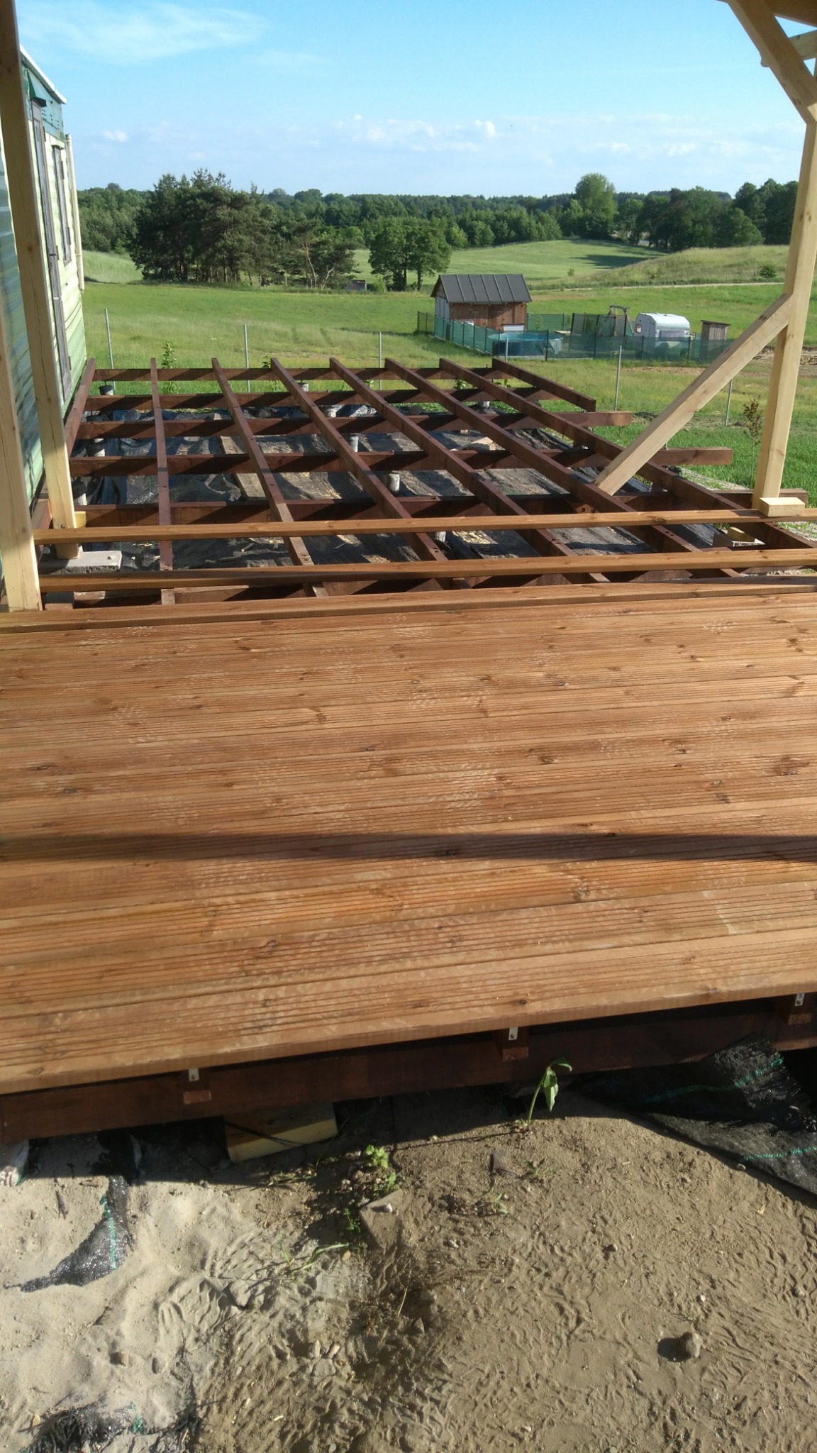 Budowa tarasu drewnianego: widok na częściowo ułożone deski i konstrukcję nośną na tle zielonego krajobrazu z budynkiem gospodarczym w oddali.