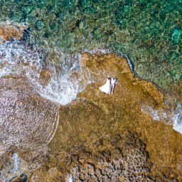 Tomasz Michalak - Fotografia - Widok z góry na parę leżącą na skalistym brzegu obok turkusowej wody morskiej, panna młoda w białej sukni.