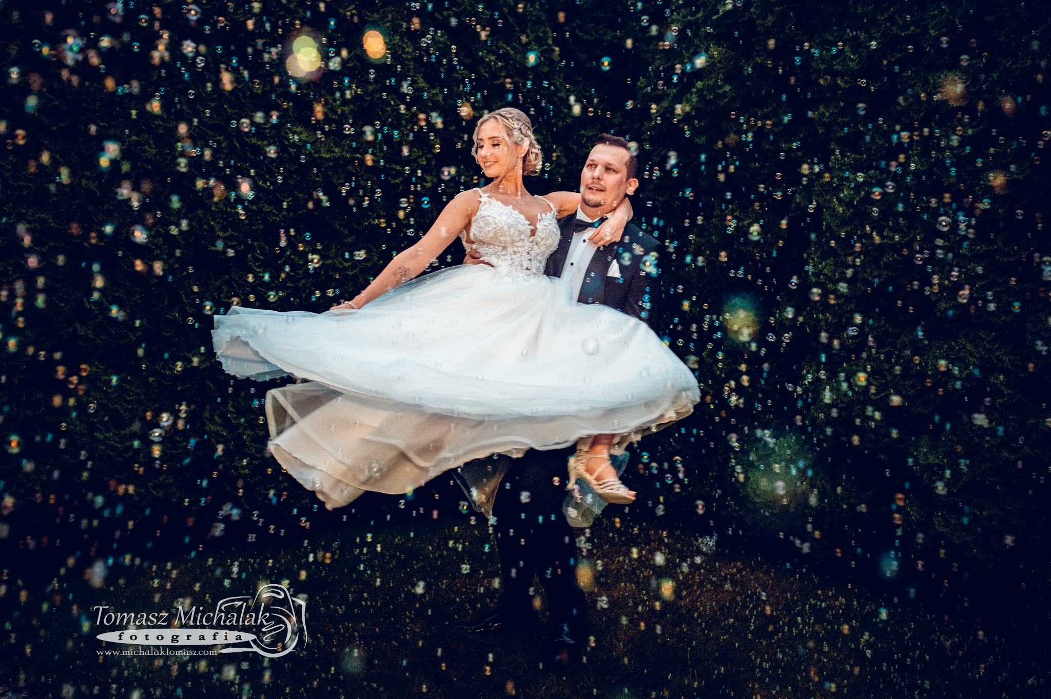 Uśmiechnięta para młoda w tańcu, otoczona bańkami mydlanymi na tle ciemnej zieleni.