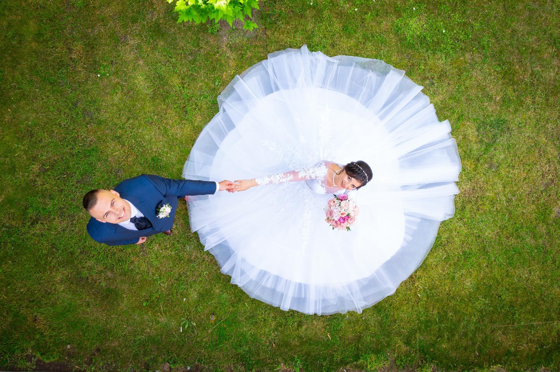 Panna młoda w białej sukni z bukietem różowych kwiatów i pan młody w granatowym garniturze, trzymający się za ręce, sfotografowani z góry na zielonym trawniku.