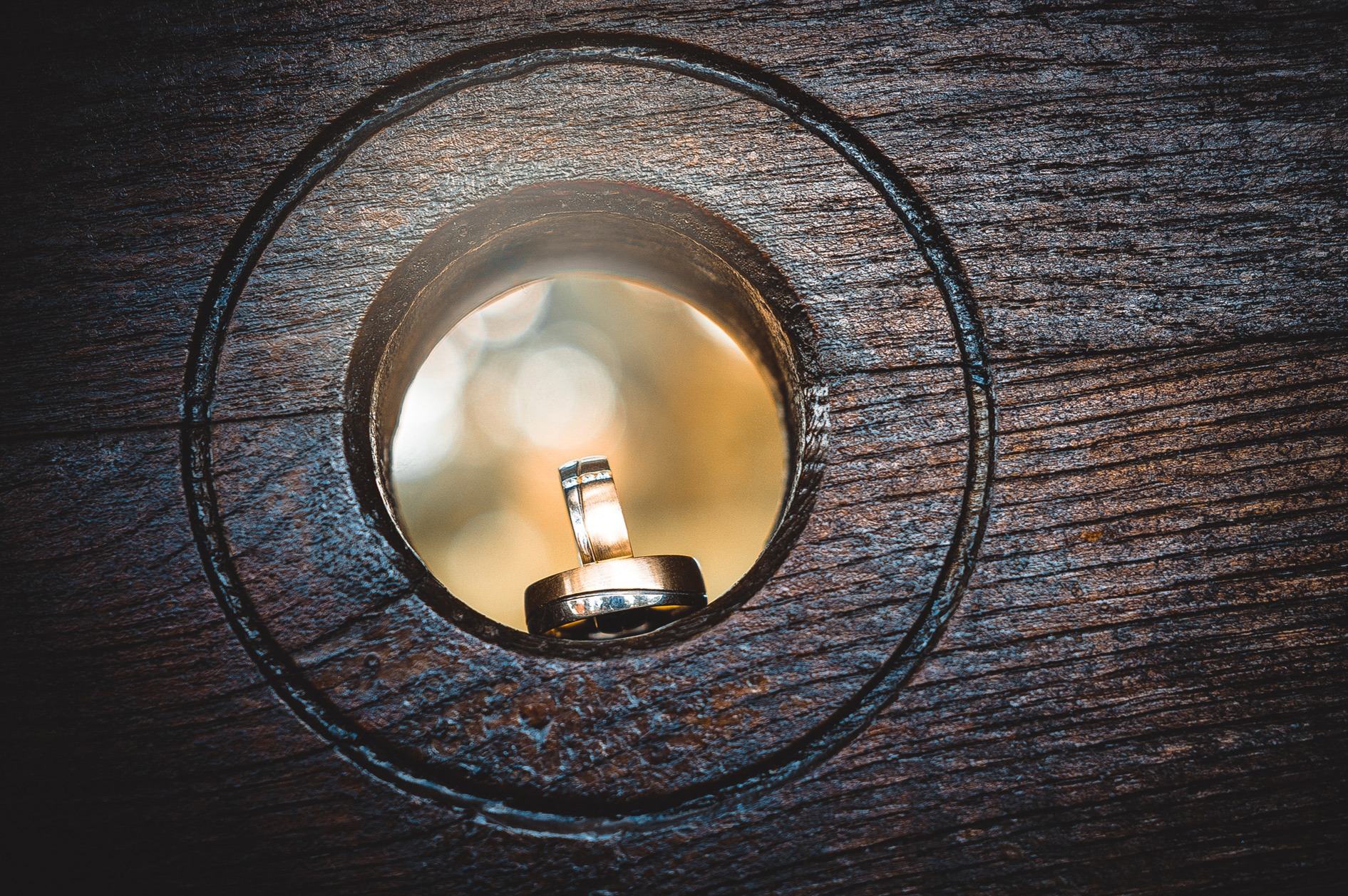 Dwa złote obrączki ślubne umieszczone w okrągłym otworze w ciemnym, teksturowanym drewnie, z rozmytym, ciepłym tłem.