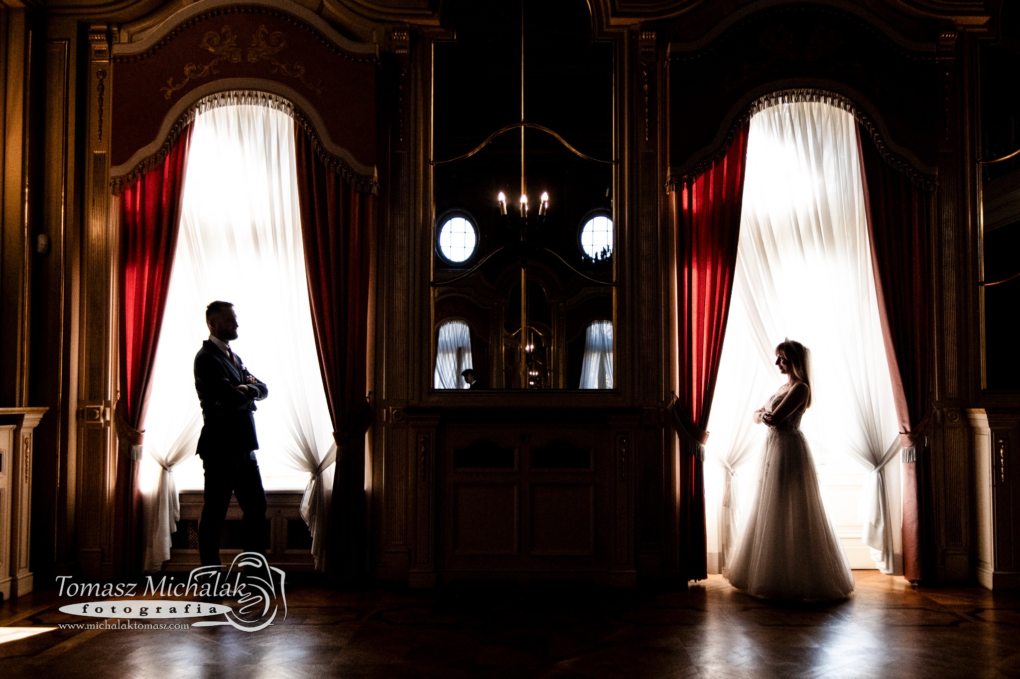 Silhouetted para młoda stoi w oknach z czerwonymi zasłonami w eleganckim wnętrzu pałacu, odbicie żyrandola w lustrze.