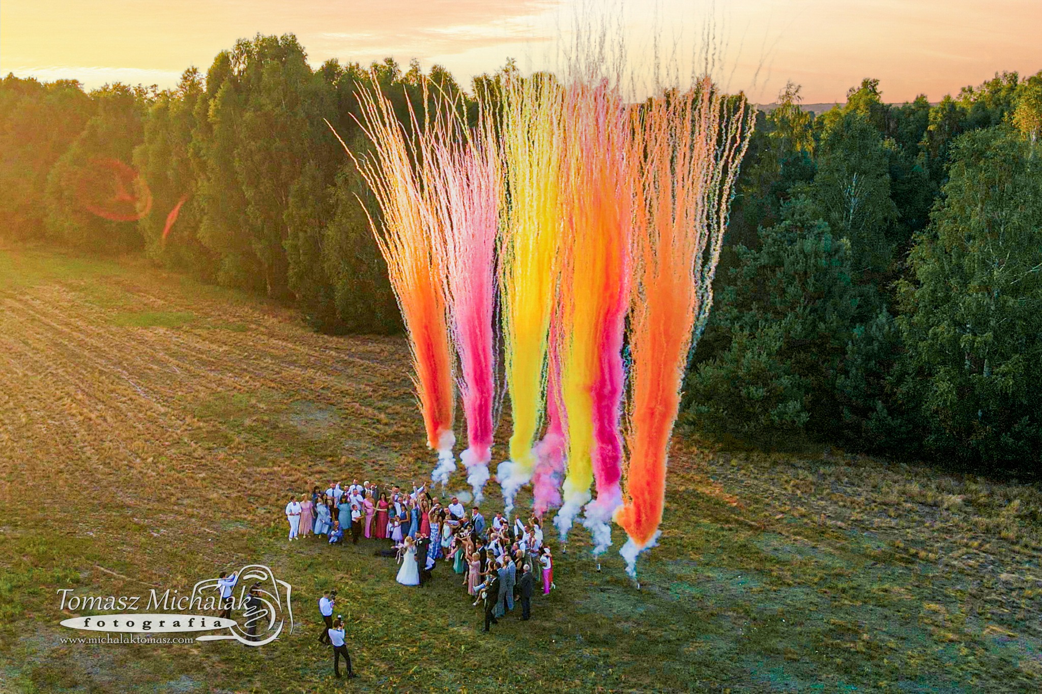 Zdjęcie z drona przedstawia grupę ludzi, prawdopodobnie nowożeńców i gości, stojących na łące w otoczeniu kolorowych, pirotechnicznych fontann dymnych w odcieniach różu, żółci i pomarańczy; w tle...