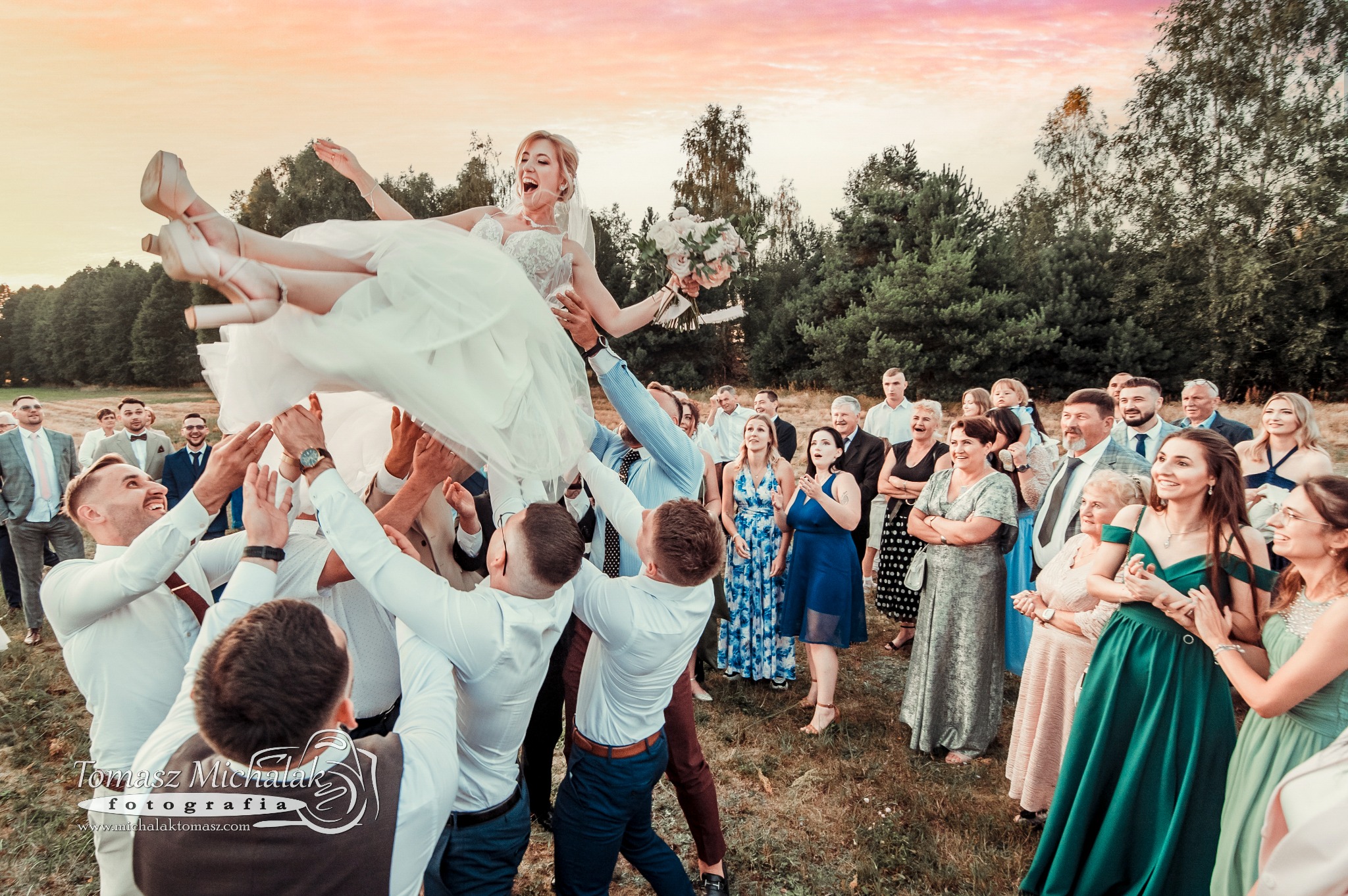 Radosna panna młoda w białej sukni i na wysokich obcasach, podrzucana przez gości weselnych na tle zachodzącego słońca i zieleni drzew.
