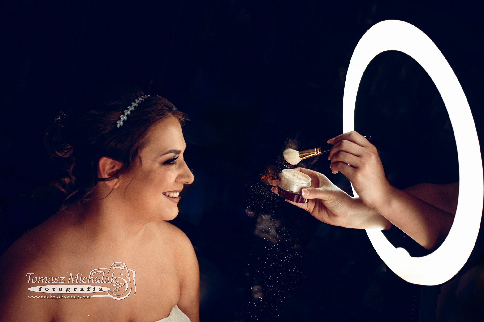 Uśmiechnięta kobieta z delikatnym diademem we włosach podczas nakładania pudru pędzlem, w tle ciemne tło i okrągła lampa doświetlająca.