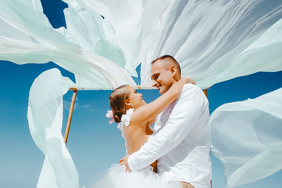 Para młoda obejmująca się pod baldachimem z powiewających, jasnych tkanin na tle błękitnego nieba, panna młoda z kwiatem we włosach, uśmiechnięci i patrzący na siebie.