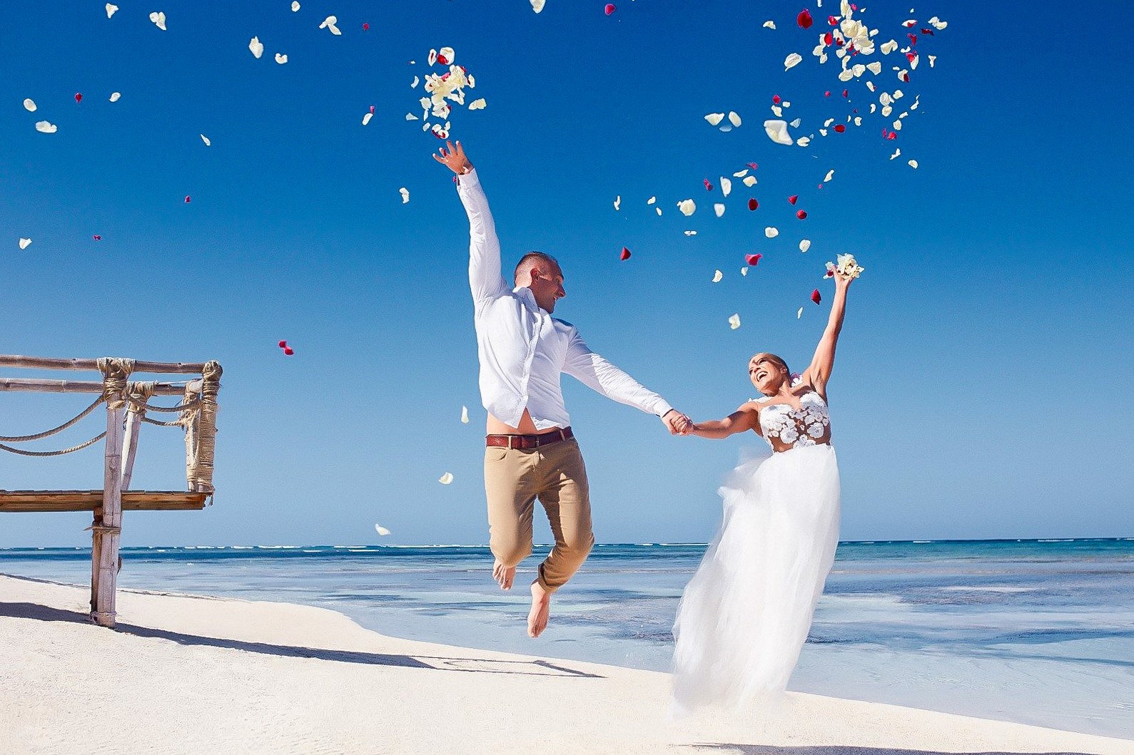 Para młoda w skoku radości na plaży, obsypywana płatkami kwiatów, błękitne niebo i morze w tle, drewniana konstrukcja po lewej.