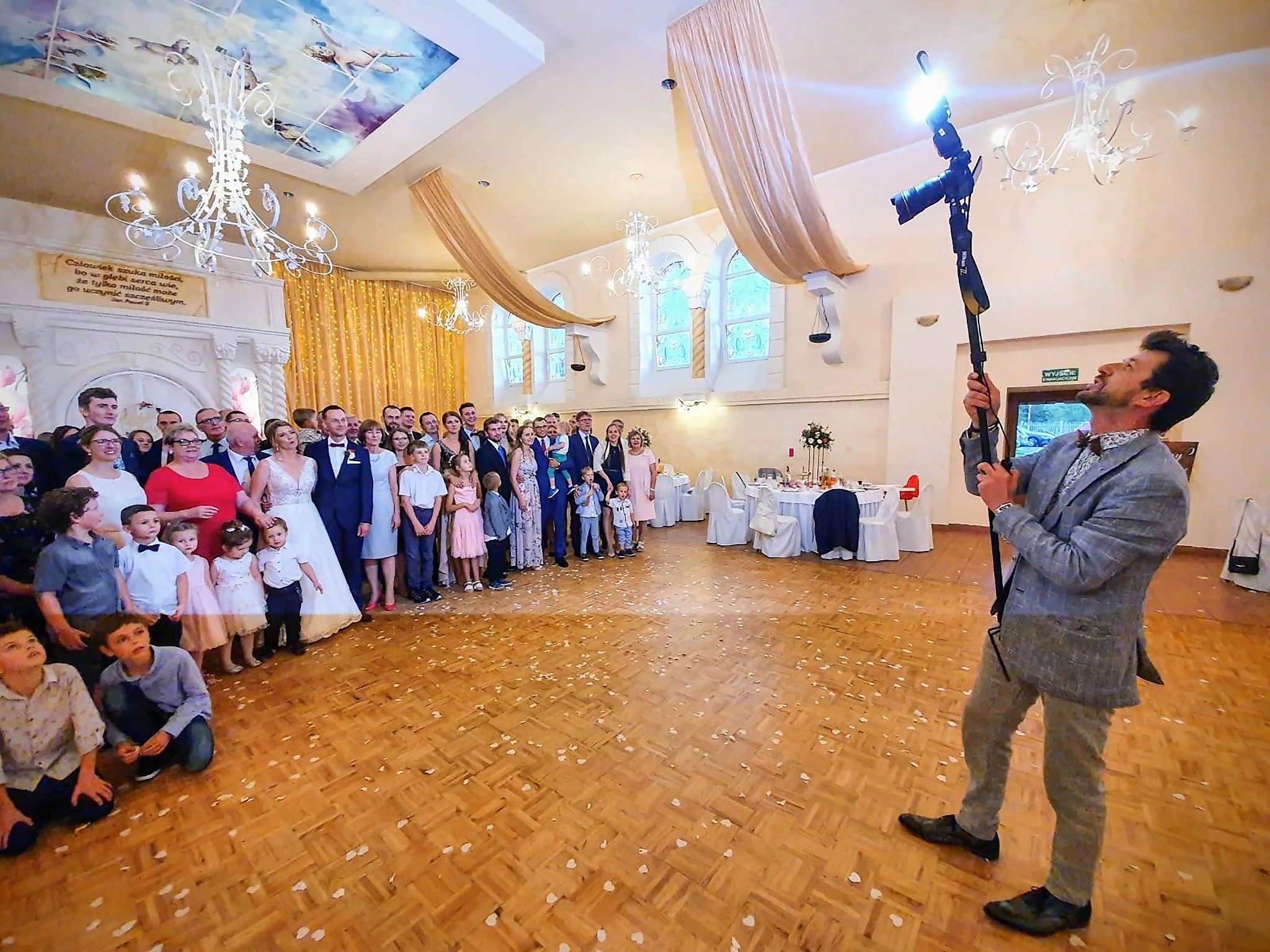 Fotograf z aparatem na wysięgniku robi zdjęcie grupie ludzi, prawdopodobnie nowożeńcom i gościom weselnym, w eleganckiej sali z parkietem posypanym płatkami kwiatów.