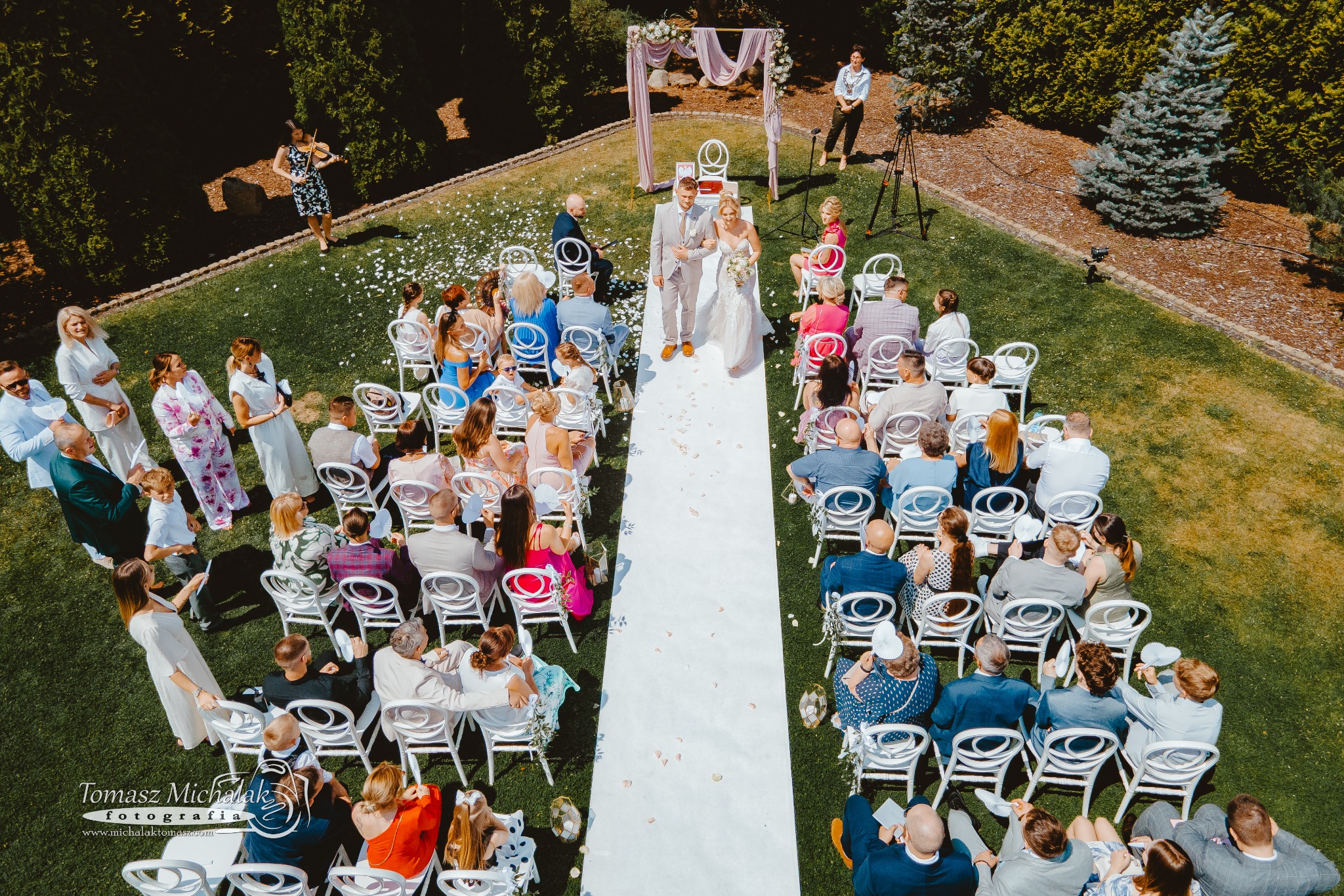Para młoda idzie białym dywanem wśród gości siedzących na białych krzesłach podczas ceremonii ślubnej w plenerze, widok z góry, w tle muzyk gra na skrzypcach.
