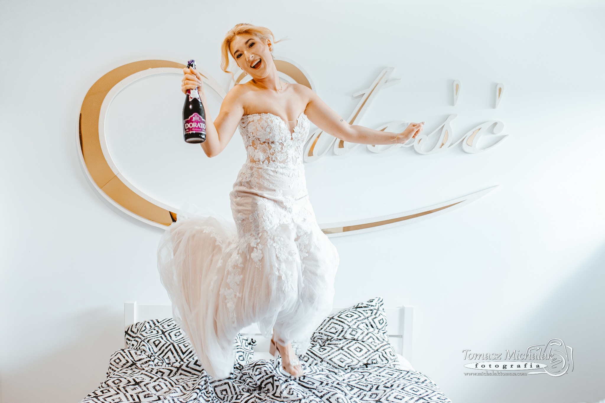 Uśmiechnięta panna młoda w koronkowej sukni, skacze na łóżku z butelką Dorato w dłoni, na tle białej ściany z dekoracyjnym napisem i złotym okręgiem.