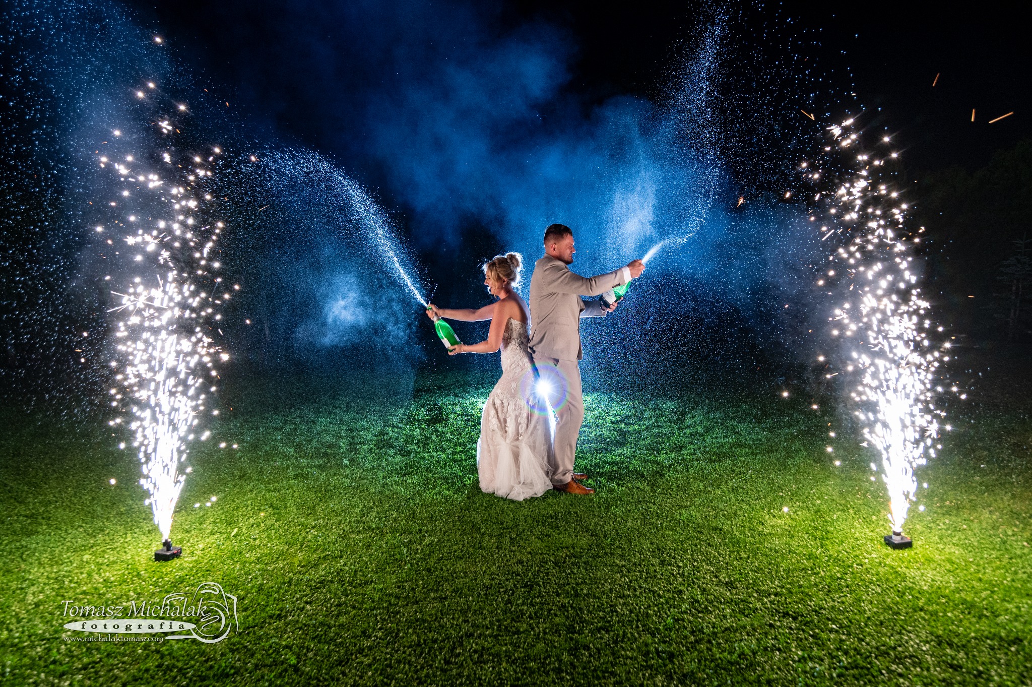 Para młoda w sukni ślubnej i garniturze otwiera butelki szampana podczas pokazu fajerwerków na trawniku w nocy.