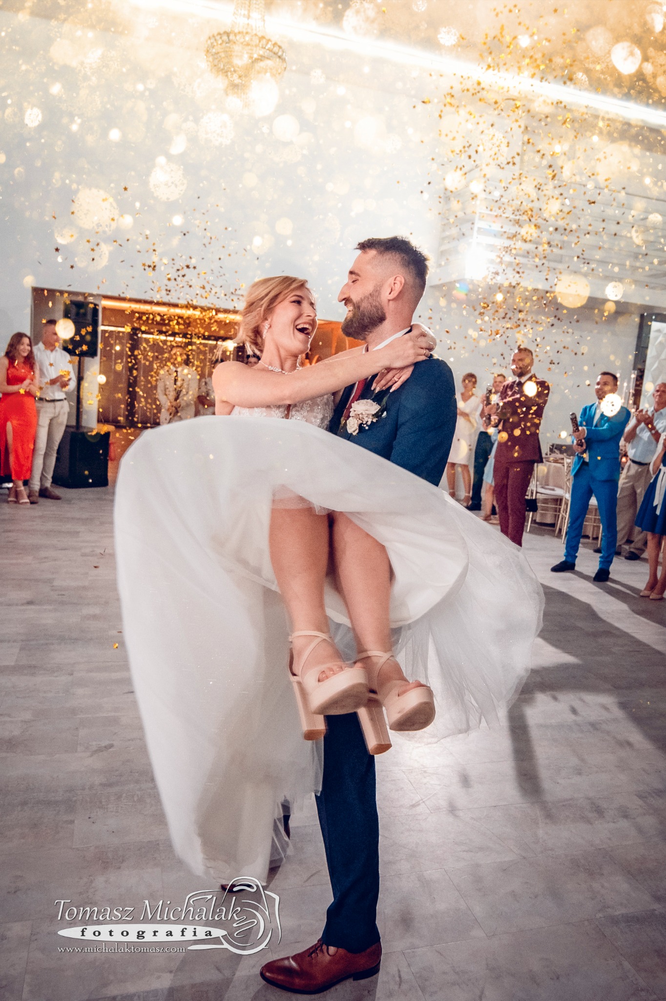 Pan młody trzyma na rękach uśmiechniętą pannę młodą podczas wesela, w tle goście i konfetti w kształcie gwiazdek.