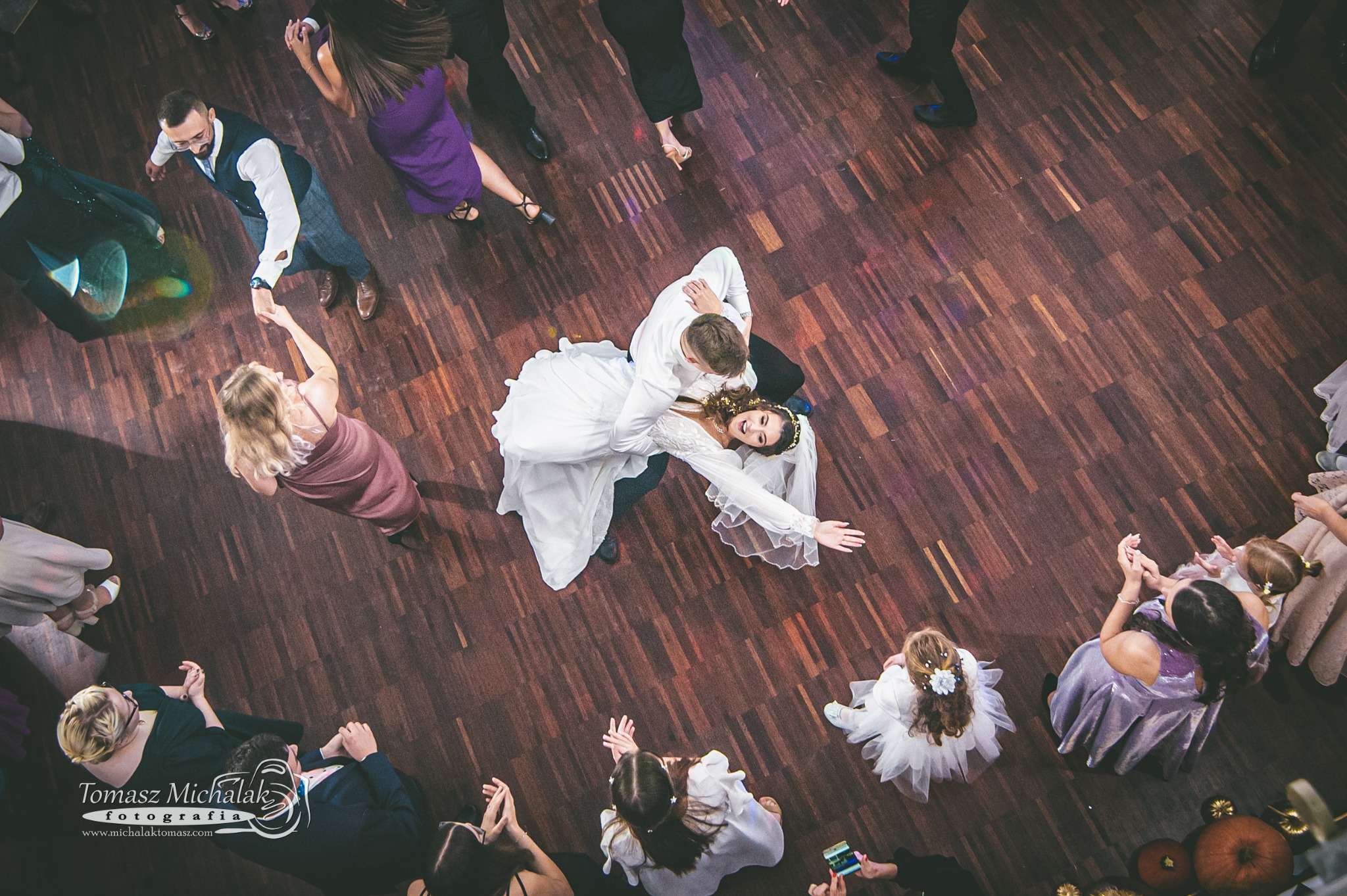 Para młoda w tańcu, widziana z góry, otoczona gośćmi weselnymi na parkiecie z ciemnego drewna.