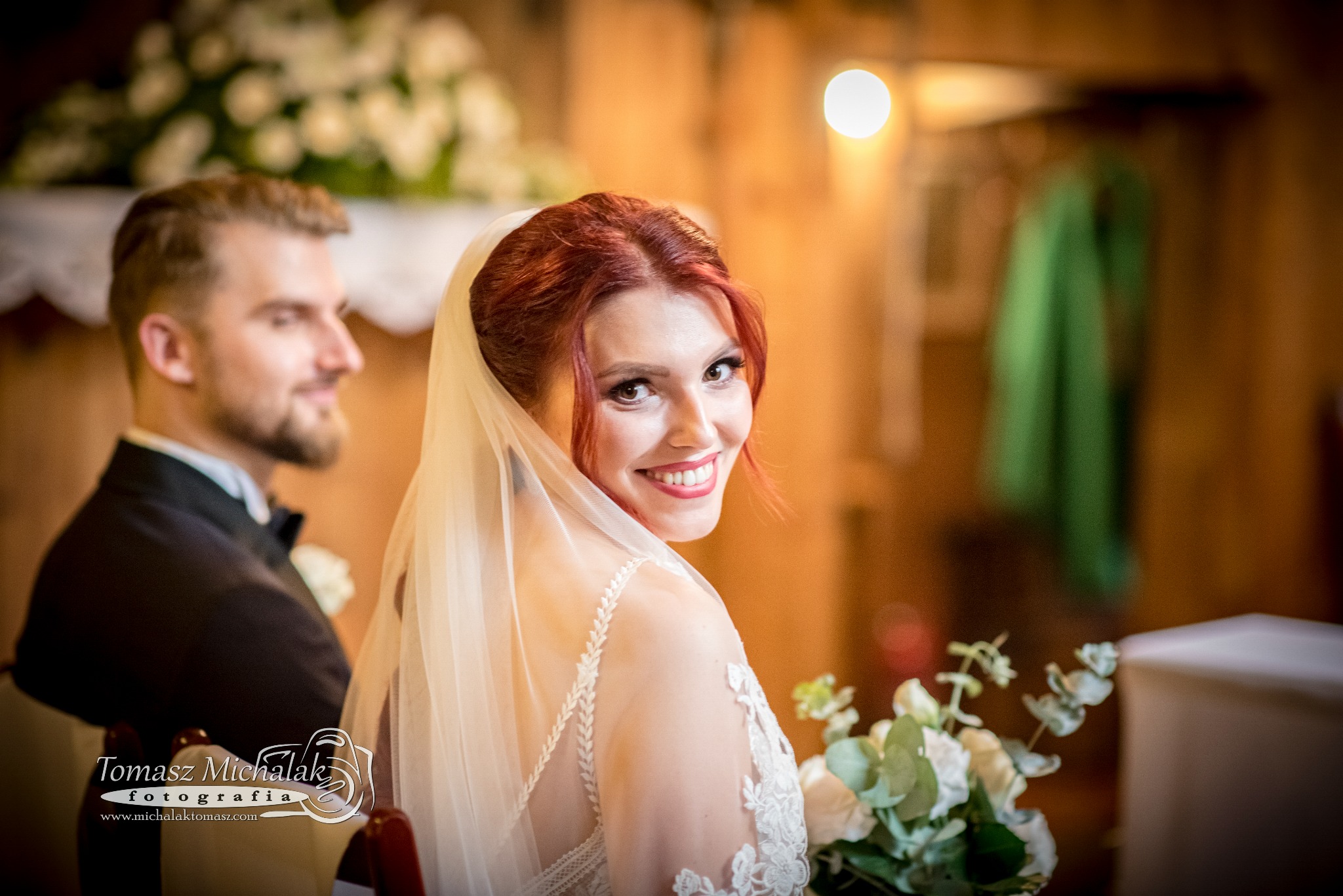 Uśmiechnięta panna młoda z rudymi włosami i welonem, odwrócona w kościele, z bukietem w ręku, obok lekko rozmazany pan młody w tle.